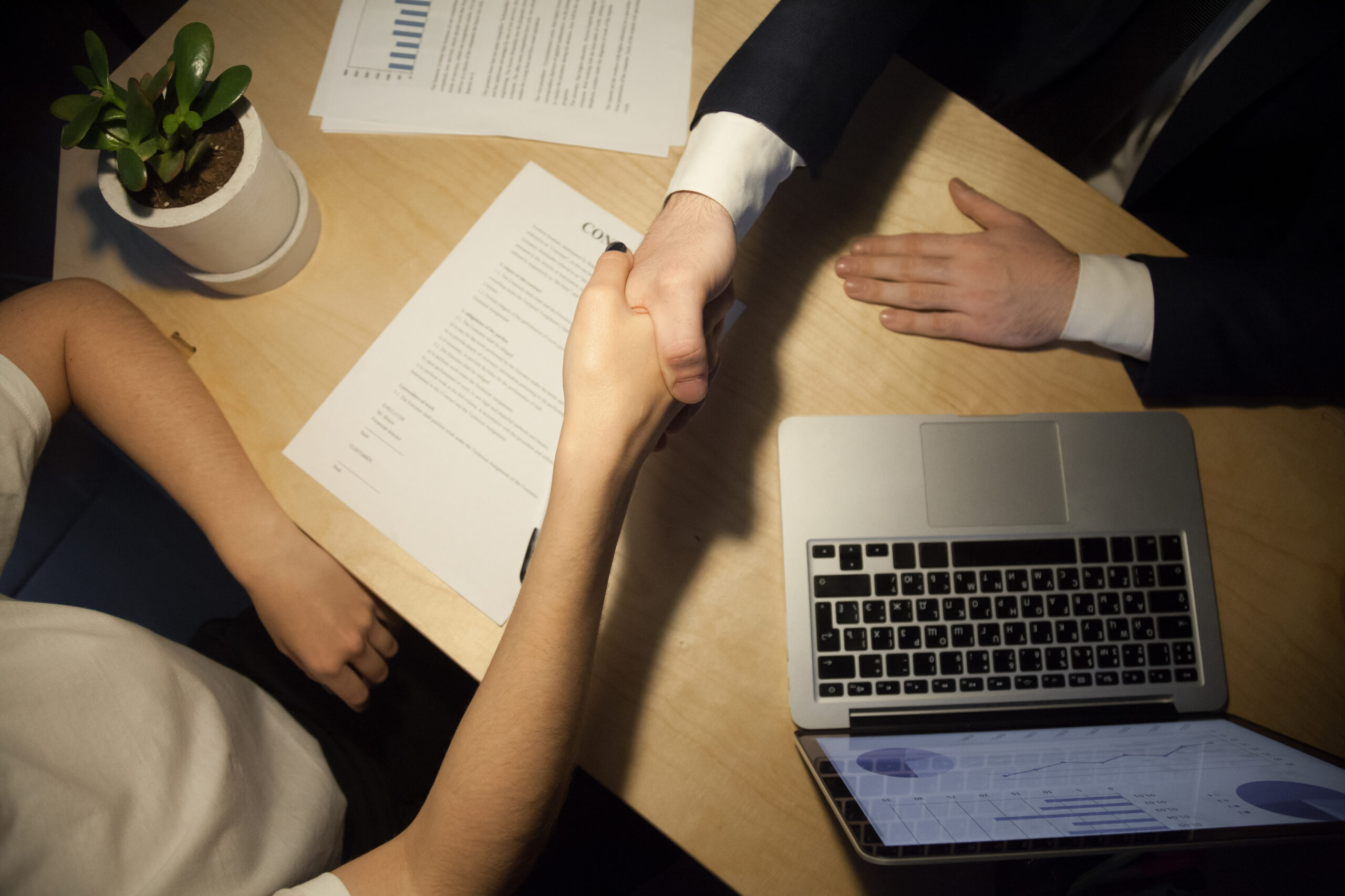 Handshake top close up view. Two businesspeople shake hands to seal a deal, laptop computer with rising stats on the desk. Human resources manager promoting successful employee. Seal a deal concept.
